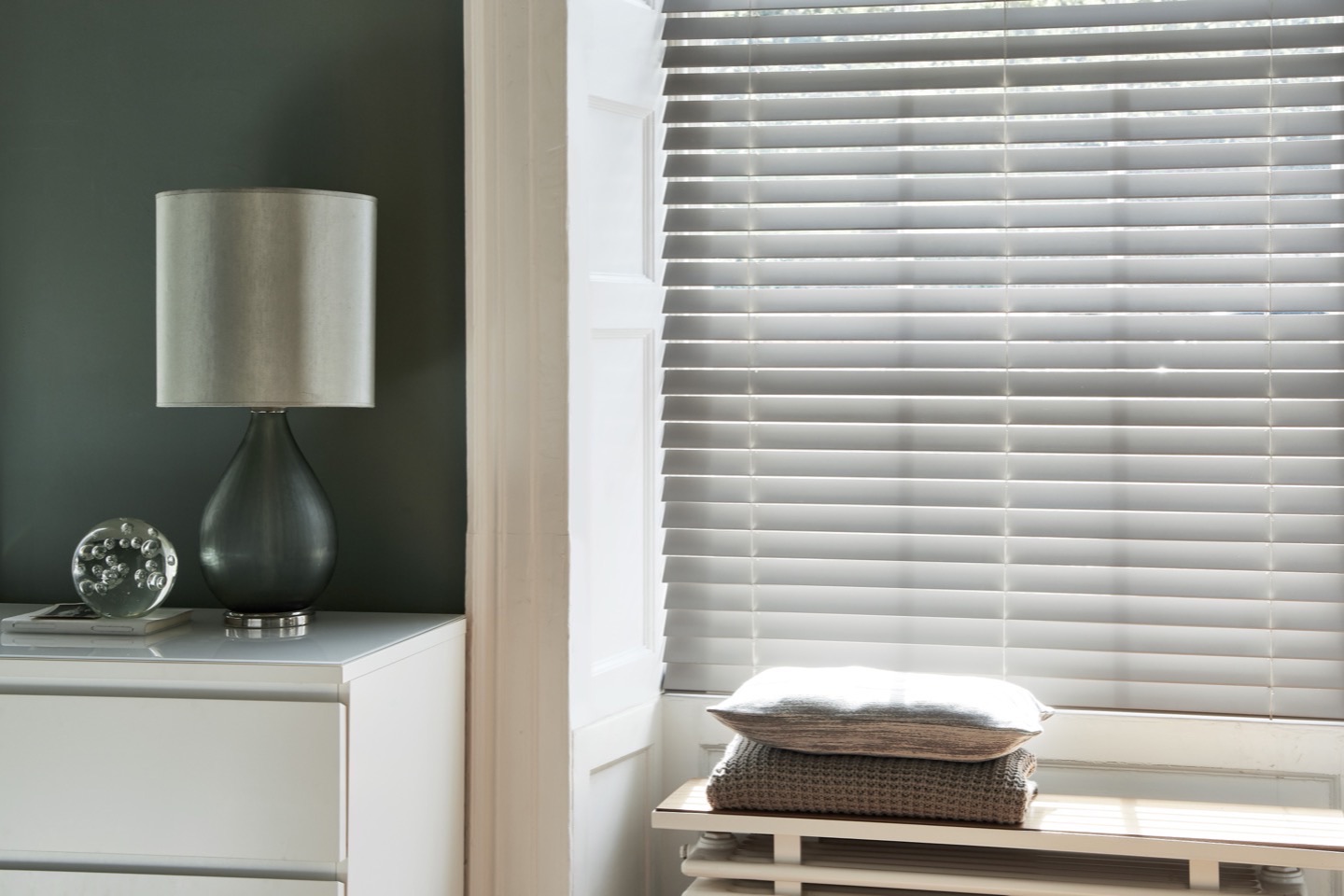 White faux wood Venetian blinds in a bedroom with a dark green wall